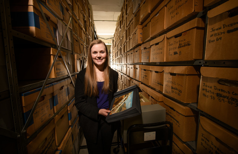 A UW student stands amongst a section of the archives at the American Heritage Center, holding a picture of a landscape.