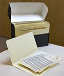 Grey archival box on table with the top lid rotated open to show the manilla file folders inside