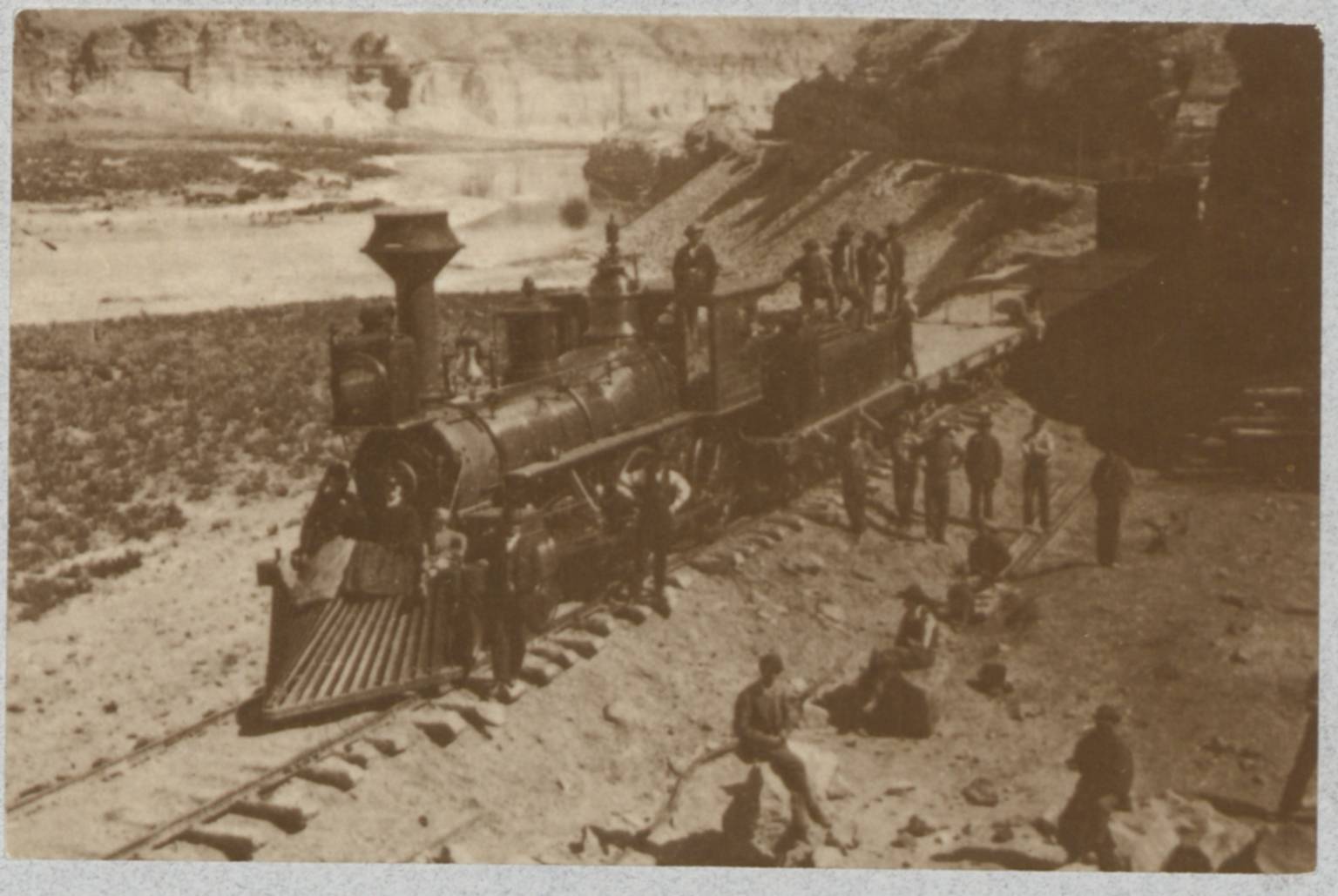 Postcard with an image of several men standing on and around a train locomotive idling on tracks by a lake in a mountain valley. From back of photo: "No. 20- Another locomotive, evidently on an excursion, at the Palisades, Green River. Photograph by Charles Savage." 