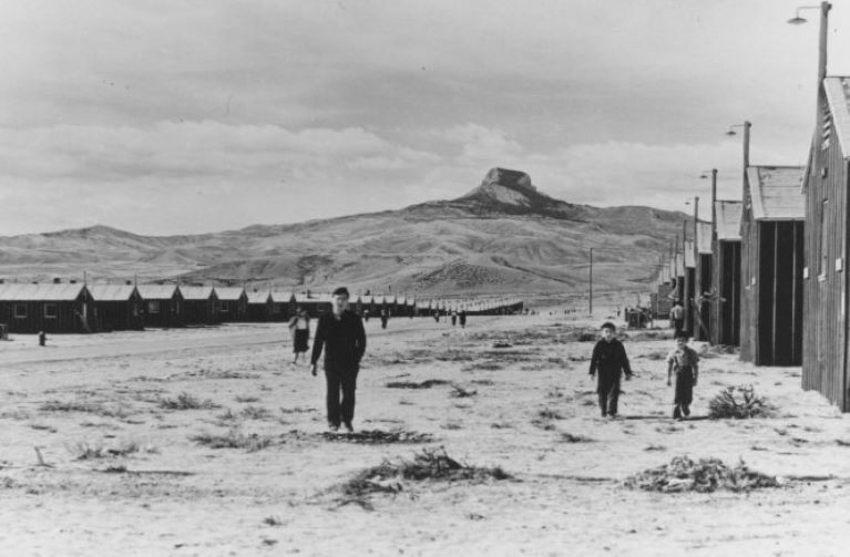 Japanese man anc children standing between rows of barracks at interment camp with heart mountain in the background