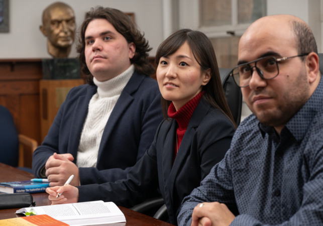 Three people sit attentively at a table with open books and notes. They appear focused and engaged in a meeting or discussion in a formal setting.