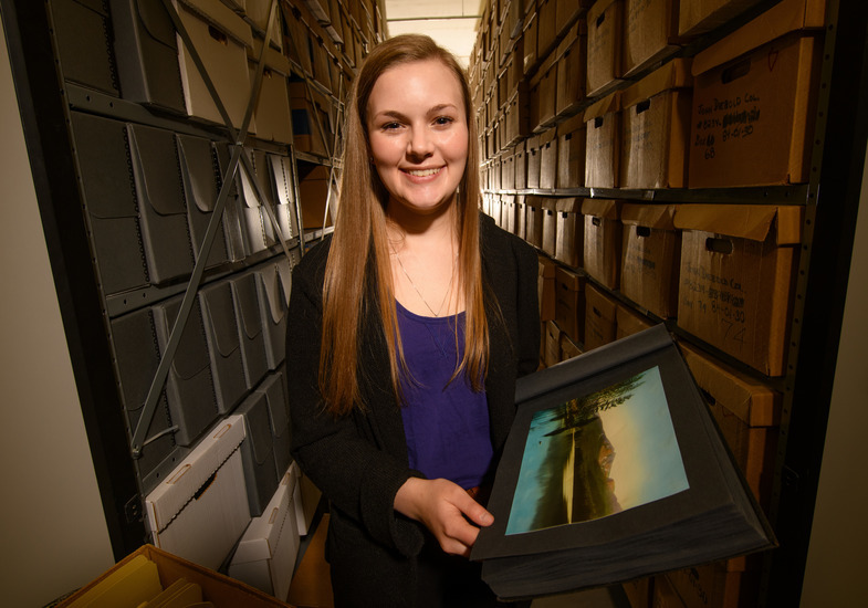 A woman stands smiling in a narrow archive with shelves of boxes. She holds an open photo album featuring a vibrant landscape image. The setting feels organized and archival.