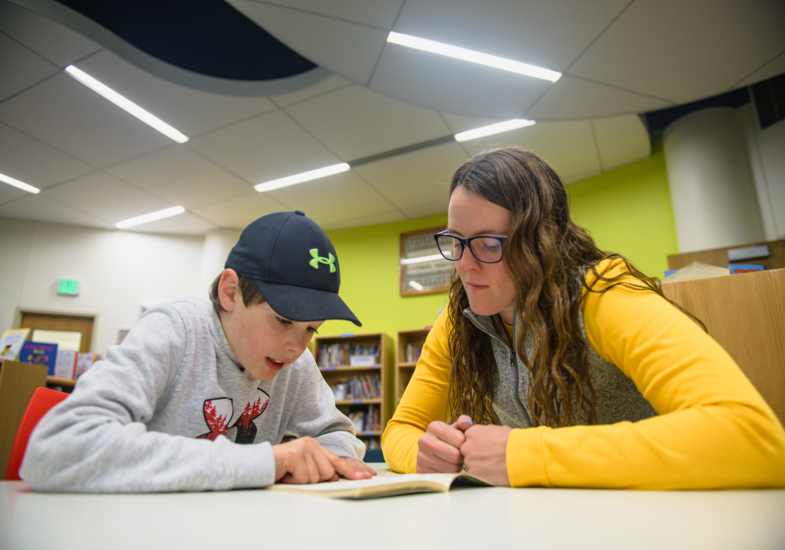 A young boy and a woman read a book together at a library table. The woman, wearing glasses and a yellow sweater, attentively listens and supports.