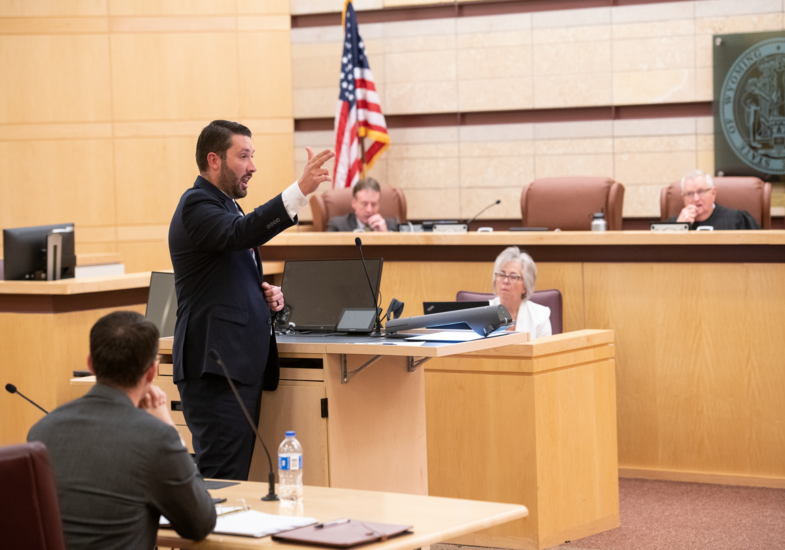 A lawyer passionately argues in a courtroom, gesturing with his hand. Seated people, a judge, and an American flag are visible in the background.