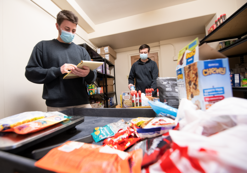 Two people in masks organize groceries in a pantry, checking items on shelves. The foreground shows cereal boxes and snack packages.