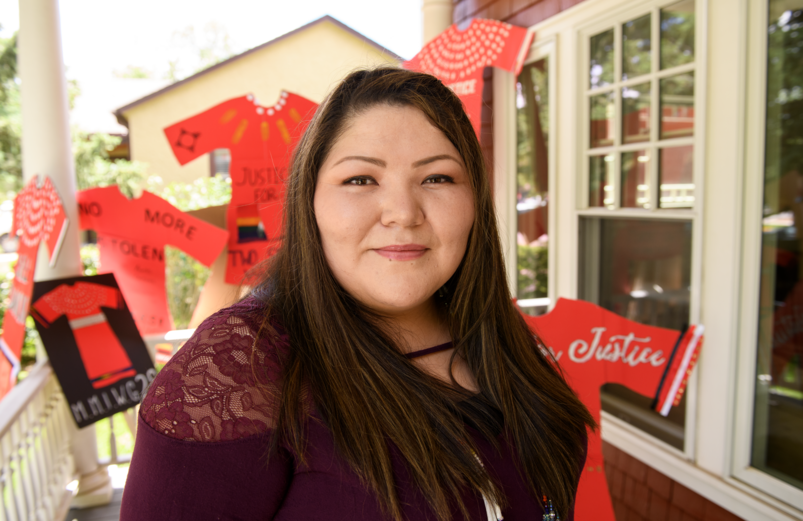 A woman in a purple top stands on a porch, surrounded by red shirts with messages like "Justice" and "No More Stolen." The scene conveys activism.