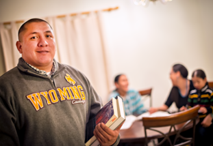 A man in a Wyoming sweatshirt holds books, smiling confidently. In the background, three people sit at a table in a warmly lit room, suggesting a family or study setting.