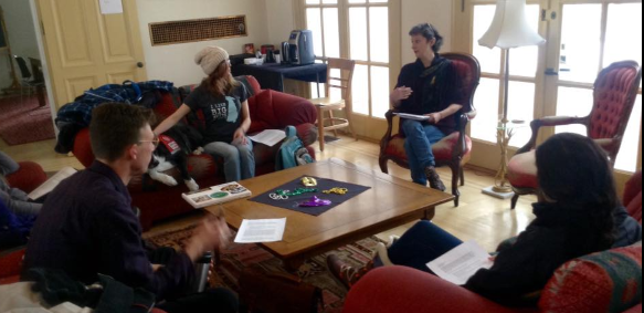 A small group of people are engaged in discussion in a cozy living room, sitting on red and brown chairs and sofas. They hold papers, creating a collaborative atmosphere.