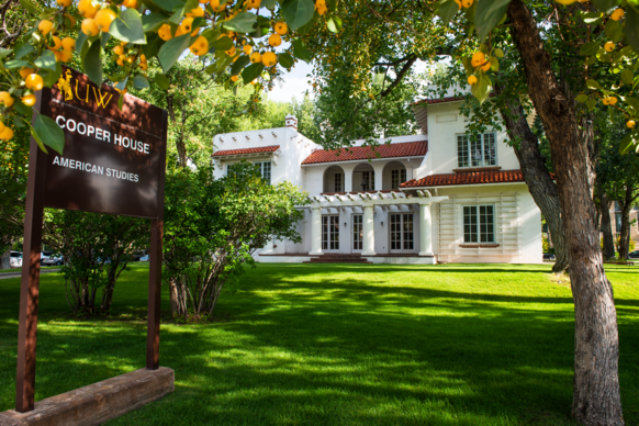 A charming, white two-story house with red-tiled roof sits among lush greenery. A sign reads "Cooper House, American Studies." Sunny and inviting atmosphere.