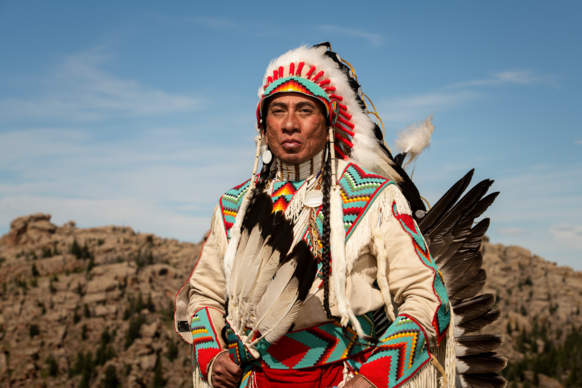 A person in traditional Native American attire stands against a rocky landscape. They wear a vibrant feathered headdress and embroidered clothing, exuding pride.