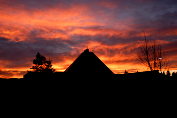 Silhouette of a building at sunset with a dramatic sky ablaze in hues of orange and purple. Trees frame the structure, creating a serene and striking scene.