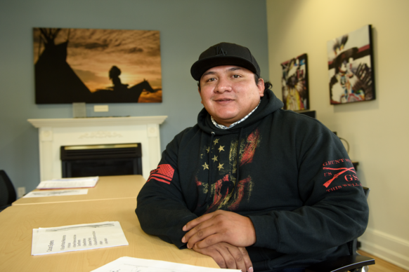A smiling person wearing a black hoodie and cap sits at a table with documents. Behind them are artwork pieces and a fireplace, creating a warm atmosphere.