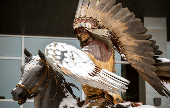 Statue of a Native American chief on a horse with large feathered wings, dusted in snow, set against a modern building backdrop, evoking a sense of strength.