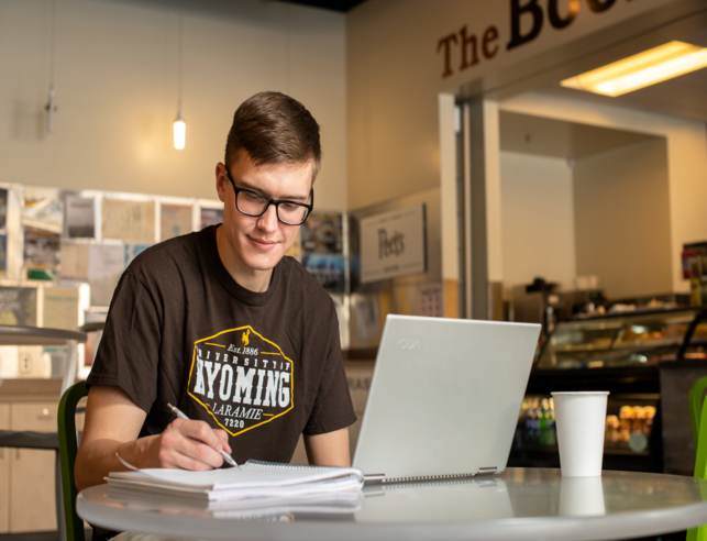 Young man in glasses studying at a cafe. He's writing in a notebook beside a laptop on a round table, reflecting focus and concentration.