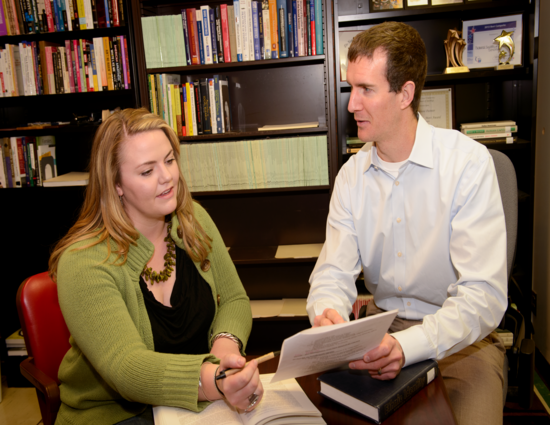 A woman in a green sweater and a man in a light blue shirt discuss documents in an office with bookshelves. The mood is collaborative and focused.