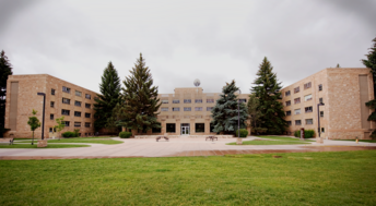 Ross Hall Ross Hall at the University of Wyoming, a large, symmetrical brick building with four stories, flanked by tall pine trees, stands under a cloudy sky, conveying a calm and serene atmosphere.