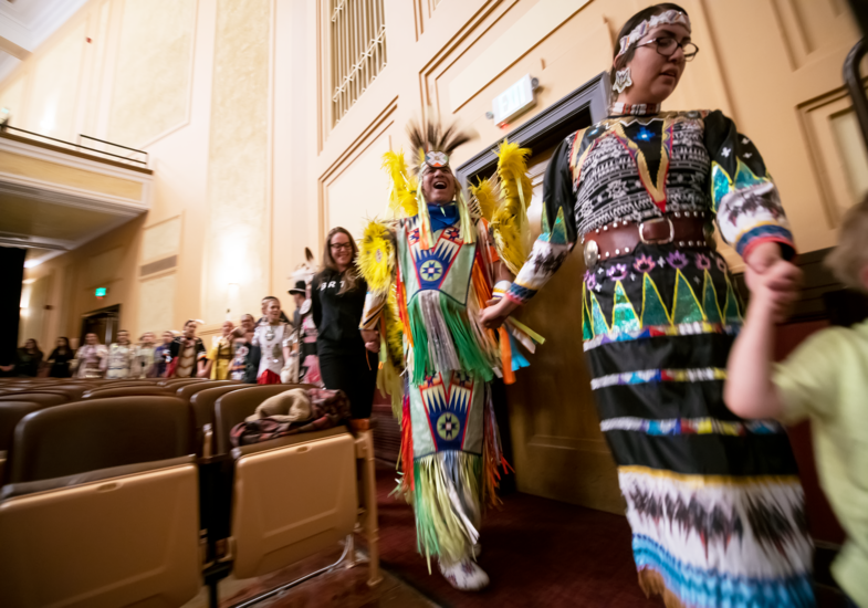 A joyful procession in a theater features people in vibrant, traditional Native American attire with colorful feathers and beadwork, exuding celebration.