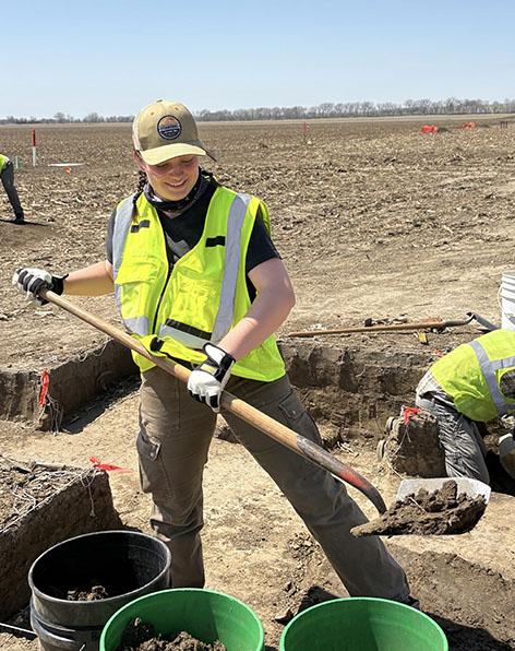 Student in a safety vest shoveling soil from an excavation trench into buckets in a field.