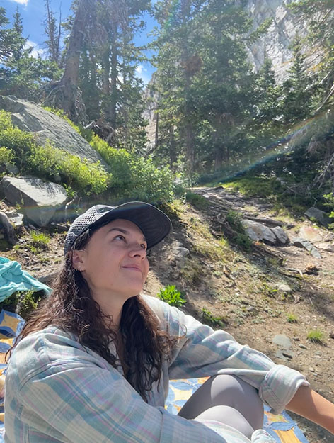 Person in a baseball cap and plaid shirt sitting on a rocky, sunlit forest trail.