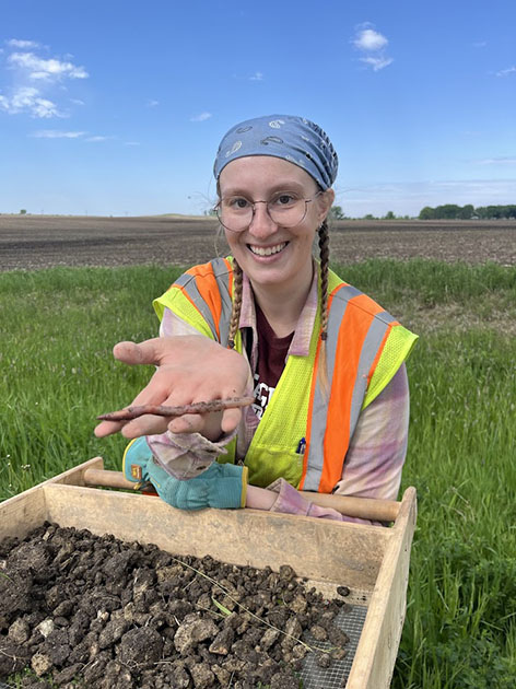 Smiling field researcher holds several earthworms over a soil-sifting tray in a grassy agricultural field.