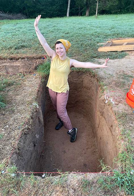 A woman stands with her arms outstretched in a freshly dug excavation pit.