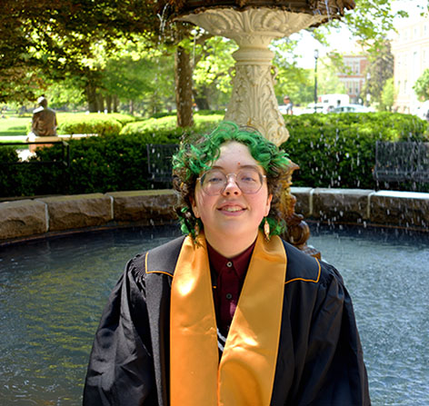 Smiling graduate in a black gown and gold stole poses in front of a large outdoor fountain.
