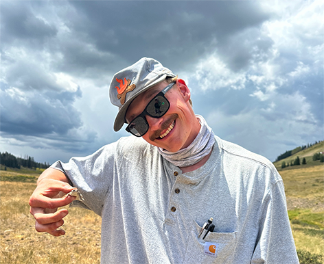 A smiling man in a hat holds a small lizard while standing in a grassy, mountainous landscape under a dramatic sky.