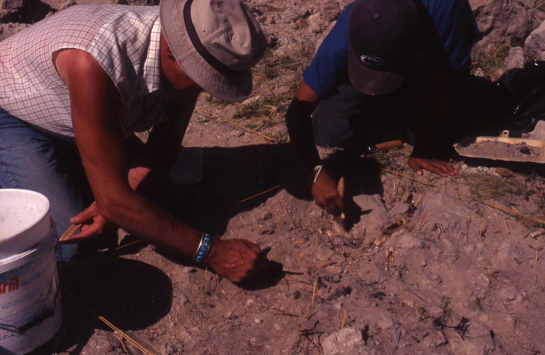 Two people are kneeling on rocky ground, carefully excavating with small tools. They appear focused and deliberate, suggesting an archaeological dig.