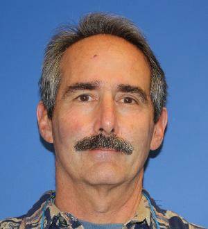 Middle-aged man with gray hair and mustache, wearing a patterned shirt, facing forward against a blue background.