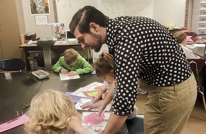 A student teacher in a black and white shirt works with art students.