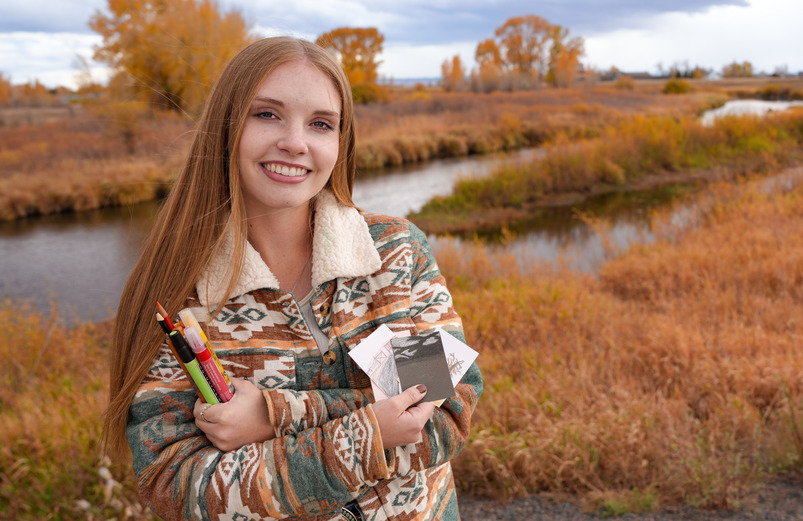 An art student smiles for a photo by a river while holding art supplies