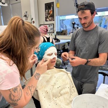 Two art students setting plaster on another student's face to make a mask during mask making workshop