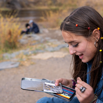 Student sketching along gravel bank of river using colored pencils