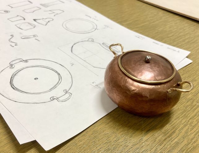 Bronze pot sitting on table near pencil blueprints of the pot