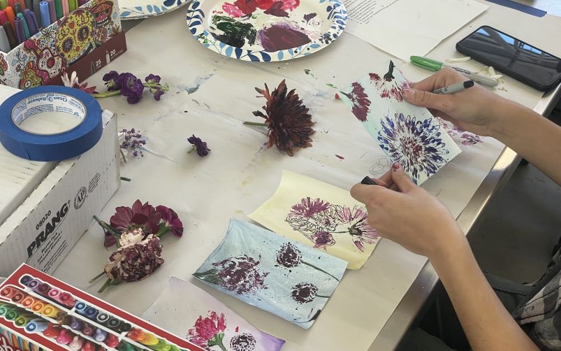 Art student holding flora drawing over a table with loose flowers, drawings, and art supplies.