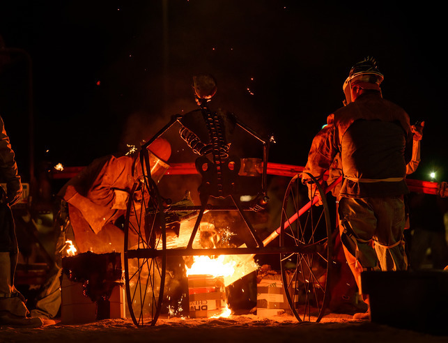 Two artists pouring molten iron, a skeleton on wheels in the foreground lit by an orange glow.