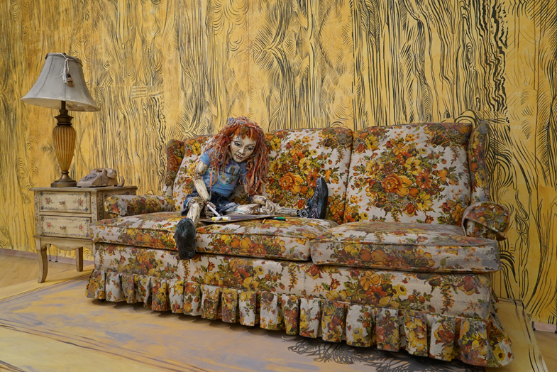 Lifesized puppet drawing with markers posed on a tacky floral couch next to a table with a gold lamp and rotery phone.