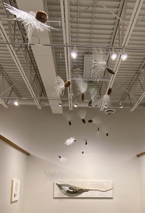 Art exhibition of dandelion seeds hanging from ceiling with effect of drifting away from dandelion sculpture mounted on far wall in background.