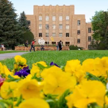 The Arts and Sciences Building on a university campus, with students walking nearby and flowers in the foreground.