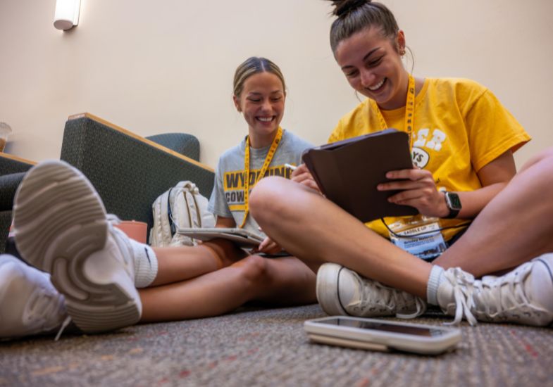Two students sit on the ground while looking and their laptops and smiling