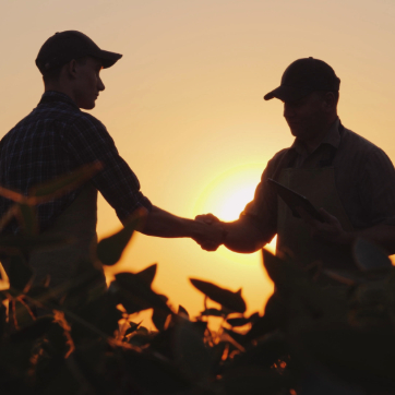 Two people in hats shaking hands in a field at sunset, silhouetted against the sun.