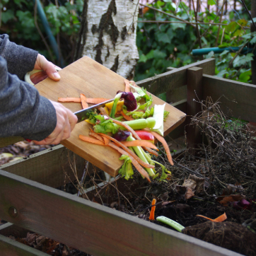 Person scraping vegetable scraps from a cutting board into a compost bin outdoors.