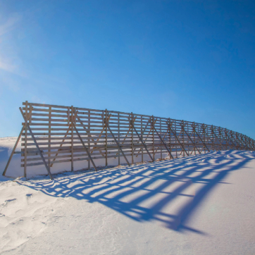 Long wooden snow fence casting shadows across a snow-covered landscape under a clear blue sky.