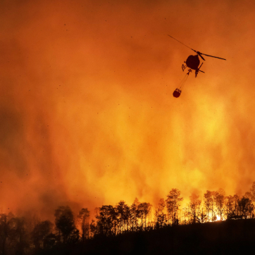 Helicopter dropping water over a forest wildfire with flames and smoke in the background.