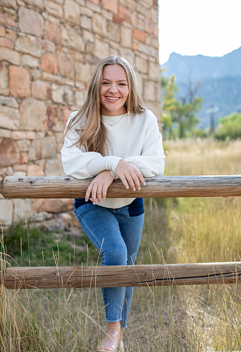 Smiling woman in a white sweater leaning on a rail fence.