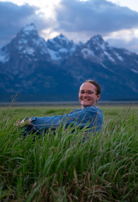 Smiling woman wearing blue sweater sitting in a mountain meadow.