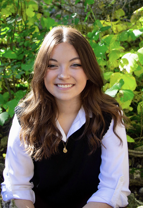 Smiling woman wearing a black vest over a white collared shirt in front of leafy vegetation.