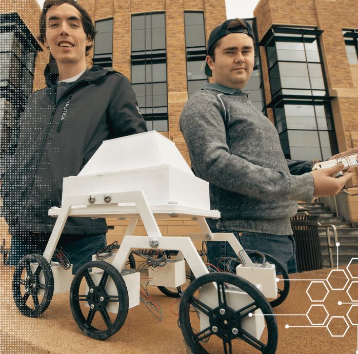 Two students pose with their robot in front of a building.