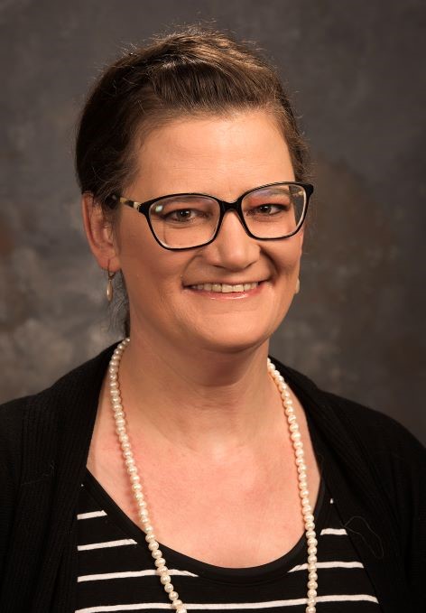Smiling woman with black shirt, black rimmed glasses, and pearl necklace