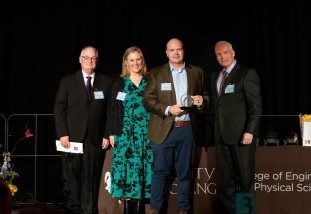 three men in suits and one woman in a dress on stage posing while handing off an award